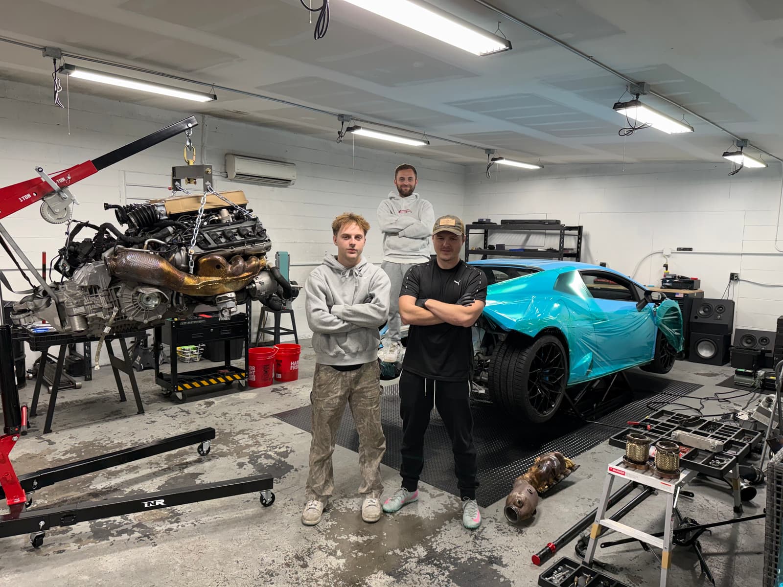 The three Rusnak brothers in the shop, arms crossed, with a V10 on the engine hoist and a wrapped Huracán on the lift behind them